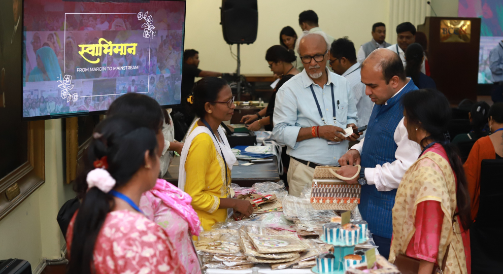 An indoor exhibition event named "Swabhiman: From Margin to Mainstream" displayed on a TV screen, where a stall presents handcrafted items like bags and pouches. A woman in a yellow kurta assists customers, including a man in a blue textured vest examining a patterned jute bag, an older man with glasses looking on, and other visitors looking at the handmade products and cupcakes on display.