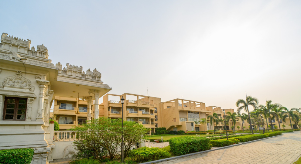 A wide exterior view of a modern senior living community in Kolkata showing residential apartments, manicured green lawns, paved walkways, palm trees, and an elegant white stone temple.