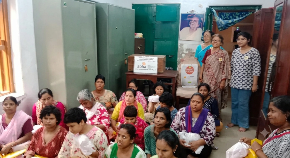 A group of women sitting together indoors during a donation drive by the ABHA Mission with organizers standing in the background.