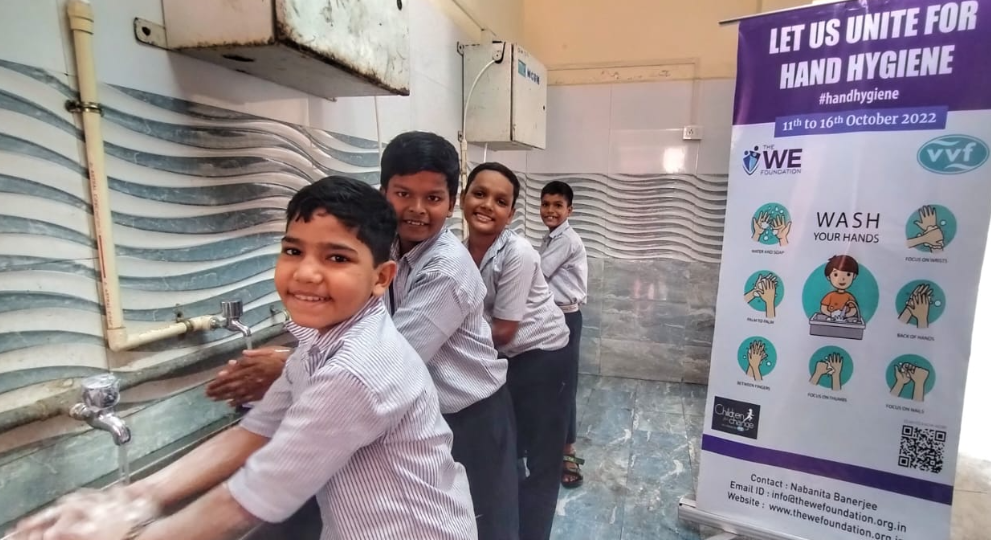 Four schoolboys smiling while washing their hands at a row of taps next to a hand hygiene awareness poster.