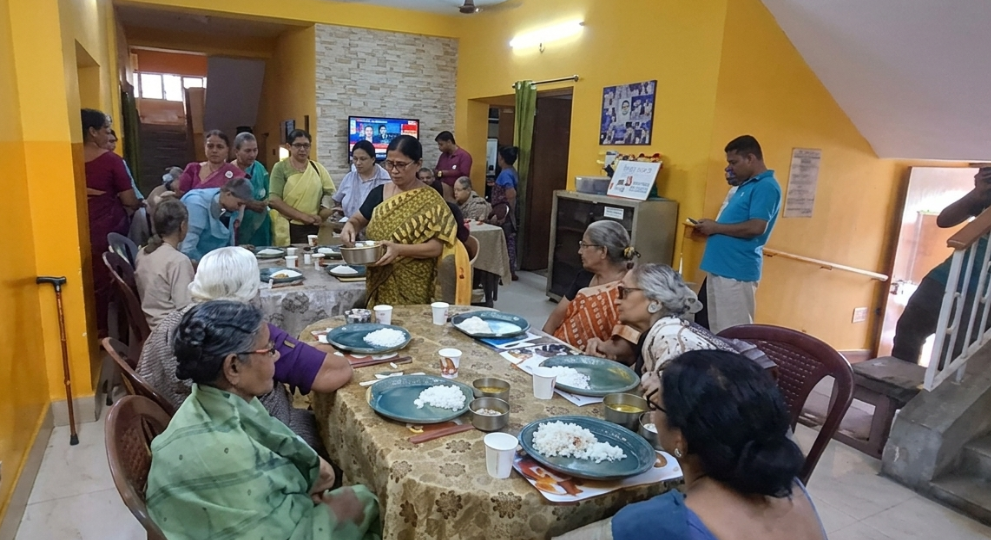 Elderly residents seated around dining tables inside a brightly painted room while volunteers serve them plated meals