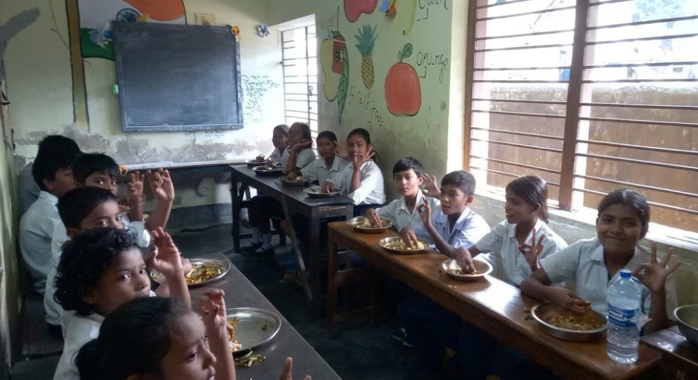 School children in white uniforms sitting at classroom desks eating meals from metal plates and making hand gestures.