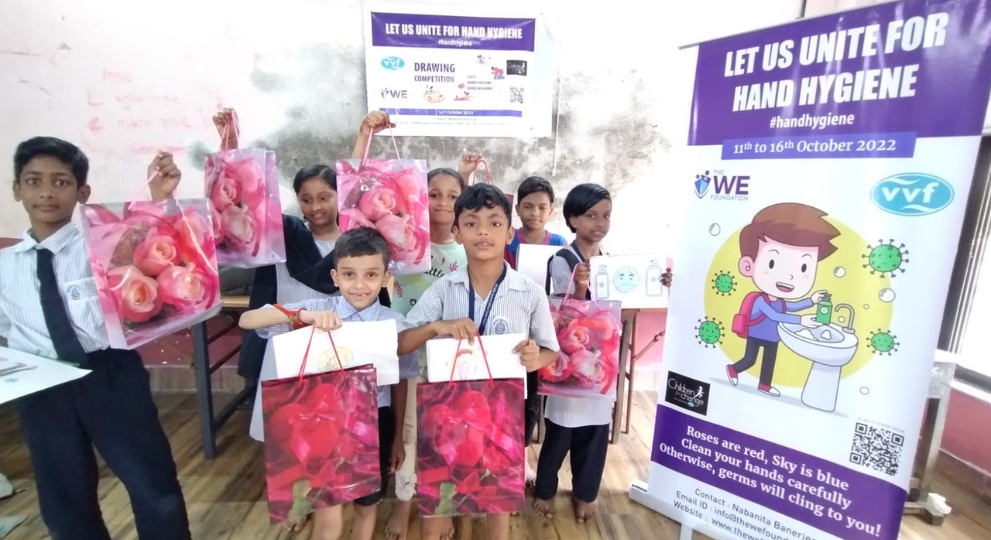 A group of smiling school children holding up pink gift bags and certificates next to a hand hygiene banner.