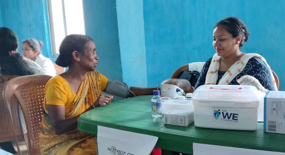 A female healthcare worker sitting at a table with a medical kit, checking the blood pressure of an elderly woman in a yellow sari.