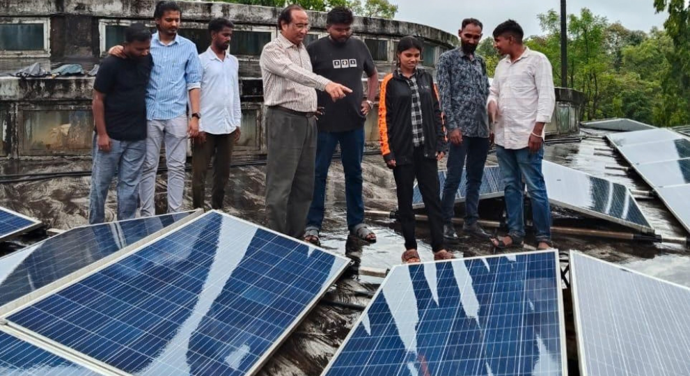 A group of people standing on a rooftop inspecting solar panels while an older man points towards one of the installations.