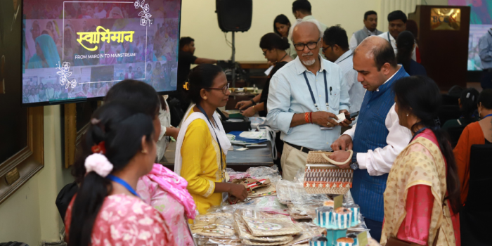 An indoor exhibition event named "Swabhiman: From Margin to Mainstream" displayed on a TV screen, where a stall presents handcrafted items like bags and pouches. A woman in a yellow kurta assists customers, including a man in a blue textured vest examining a patterned jute bag, an older man with glasses looking on, and other visitors looking at the handmade products and cupcakes on display.