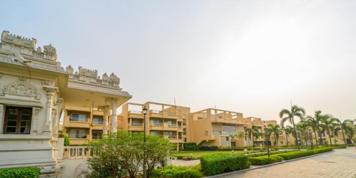 A wide exterior view of a modern senior living community in Kolkata showing residential apartments, manicured green lawns, paved walkways, palm trees, and an elegant white stone temple.