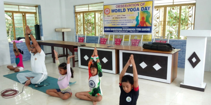 An instructor and young children sitting cross-legged on floor mats with raised overhead palms, demonstrating yoga poses during a World Yoga Day event.