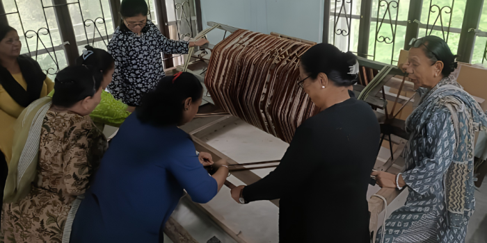 A group of women gathered around a wooden loom during a hands-on handloom weaving training session.