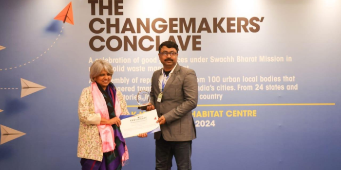 A man in a suit receiving a certificate and trophy from a woman standing on stage in front of a blue banner reading "The Changemakers' Conclave.