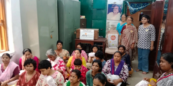 A group of women sitting together indoors during a donation drive by the ABHA Mission with organizers standing in the background.
