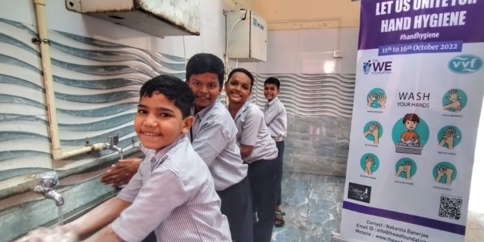 Four schoolboys smiling while washing their hands at a row of taps next to a hand hygiene awareness poster.