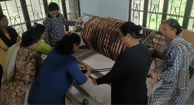 A group of women gathered around a wooden loom during a hands-on handloom weaving training session.