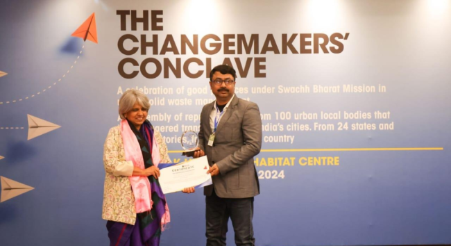 A man in a suit receiving a certificate and trophy from a woman standing on stage in front of a blue banner reading "The Changemakers' Conclave.